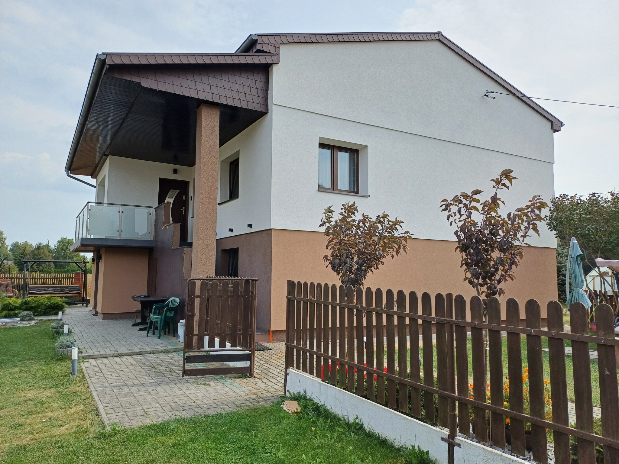 Dwukondygnacyjny dom z elewacją w odcieniach bieli i brązu, drewnianym ogrodzeniem i brukowanym podjazdem, widok z zewnątrz w pochmurny dzień.