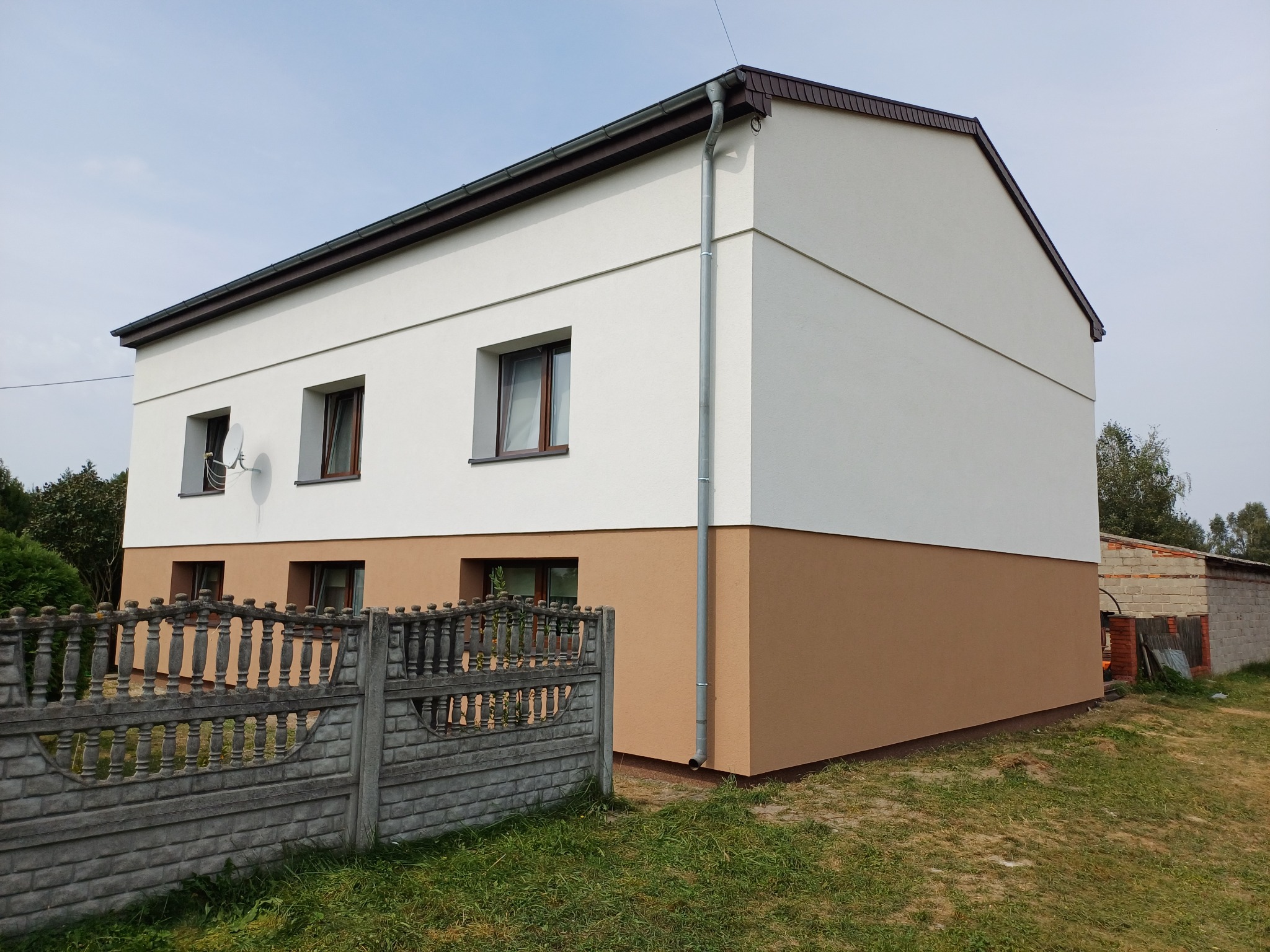 Dwukondygnacyjny dom po ociepleniu i pomalowaniu elewacji w kolorach biały i brązowy, z widocznym fragmentem betonowego ogrodzenia i satelitą na ścianie.