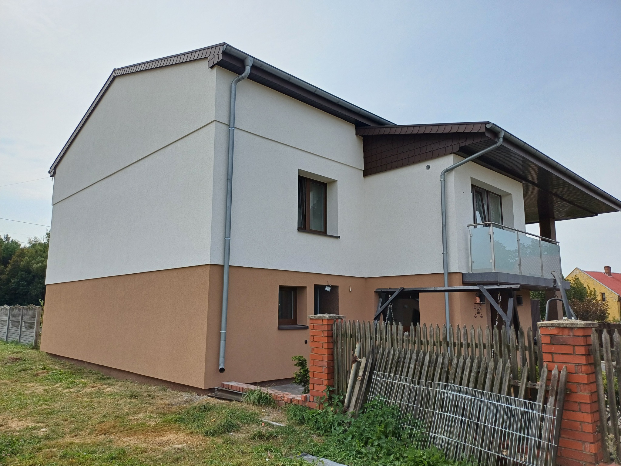 Dwukondygnacyjny dom z jasną elewacją górną i brązową dolną, z balkonem i drewnianą stolarką okienną, otoczony częściowo ceglanym i drewnianym ogrodzeniem.