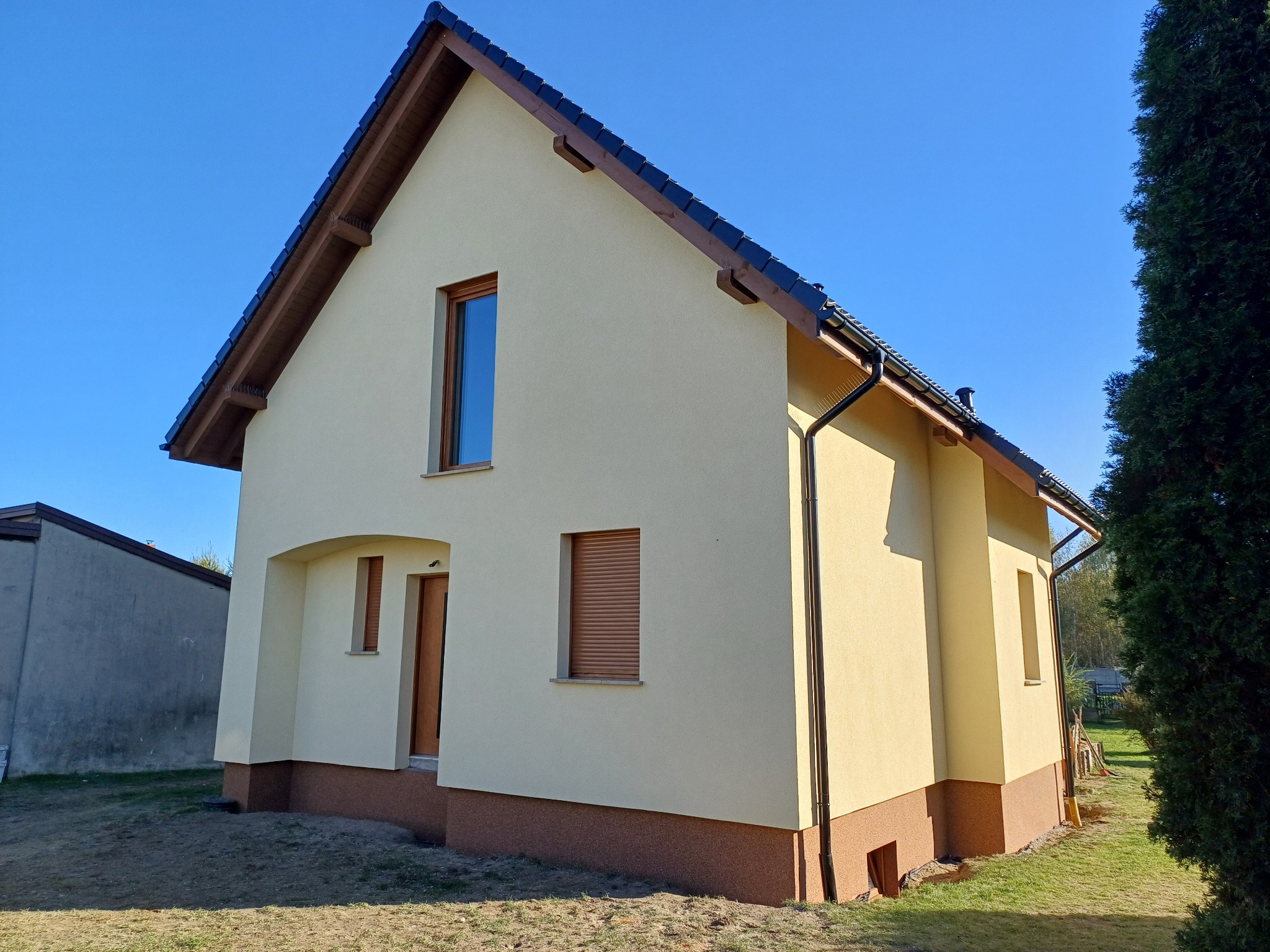 Dom jednorodzinny po remoncie elewacji, z jasną, kremową fasadą i brązowym cokołem, widoczny z perspektywy narożnej, z oknami z brązowymi roletami i ciemnym dachem na tle błękitnego nieba,...