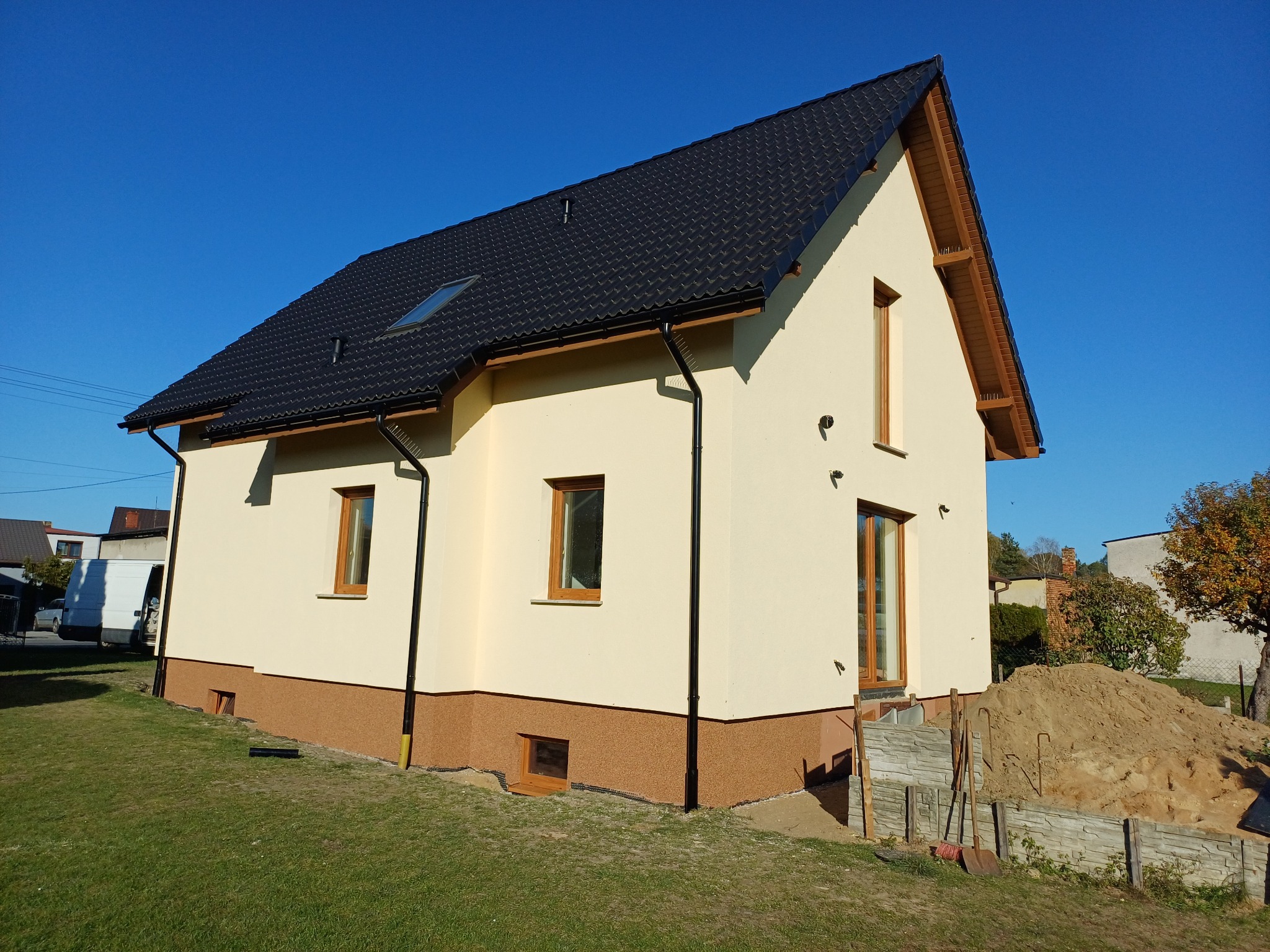 Wykończony dom jednorodzinny z beżową elewacją i ciemnobrązową podmurówką, czarną dachówką i drewnianymi oknami, widok z boku na trawnik i niebieskie niebo w tle, z widocznymi narzędziami...