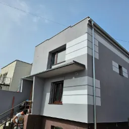 Dwukondygnacyjny dom po wykonaniu elewacji w odcieniach szarości z poziomymi pasami. Na schodach pracownik z narzędziem.