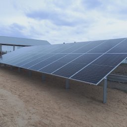 ecoVolt - Instalacja fotowoltaiczna na gruncie, panele zamontowane na metalowych słupkach, w tle budynek gospodarczy i ogrodzenie. Widok z dołu, pochmurne niebo.