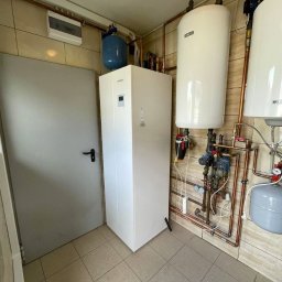 ecoVolt - Biała pompa ciepła marki Kaisai w pomieszczeniu z beżowymi płytkami, obok szare drzwi i zbiorniki wodne z miedzianymi rurami. Widok z niskiego kąta.