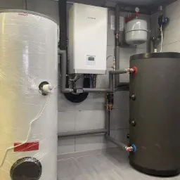 Kotłownia z dwoma zbiornikami, białym kotłem Kaisai i szarymi rurami, całość w pomieszczeniu wyłożonym jasnymi płytkami. Jeden zbiornik owinięty folią.