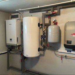 ecoVolt - Kompletna instalacja grzewcza w kotłowni: pompa ciepła Kaisai, zasobnik Galmet, naczynie przeponowe i falownik Fronius Symo. Szare rury i białe urządzenia na tle szarej ściany.