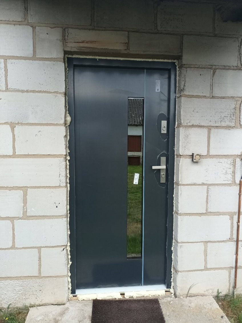 Szare drzwi wejściowe z przeszkleniem, zamontowane w surowej ścianie z betonu komórkowego. Widoczne ślady pianki montażowej.