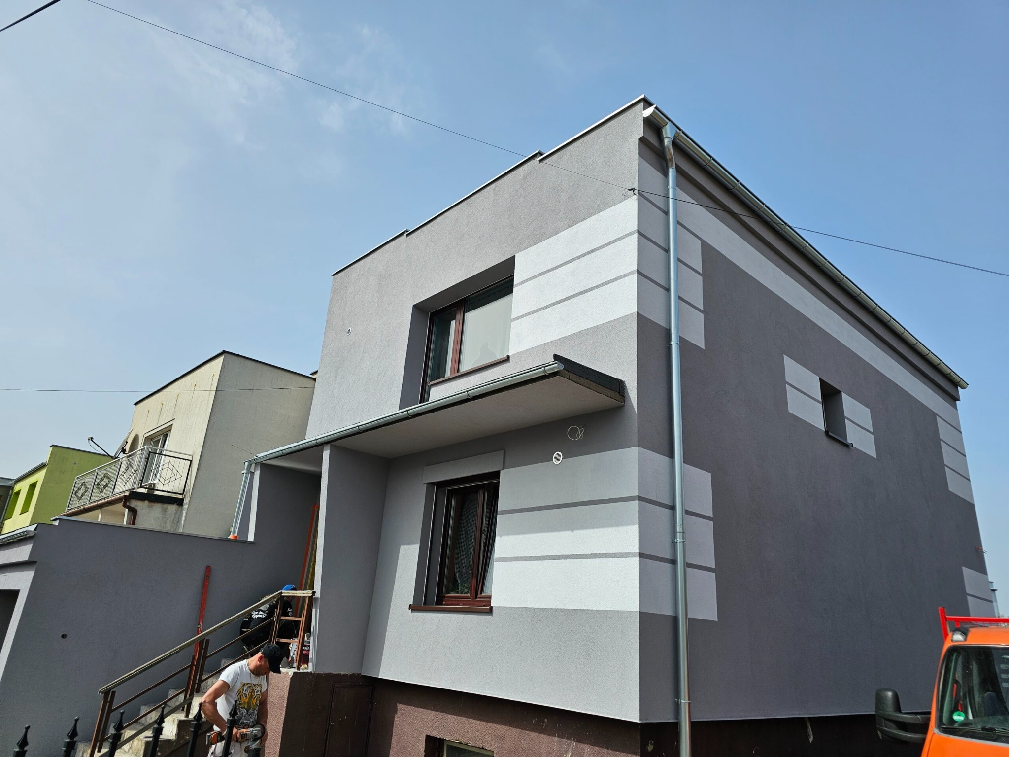 Dwukondygnacyjny dom po wykonaniu elewacji w odcieniach szarości z poziomymi pasami. Na schodach pracownik z narzędziem.