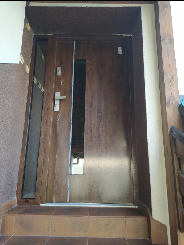 Brązowe drzwi wejściowe z szybą boczną i małym okienkiem, zamontowane na zewnątrz budynku, widoczne płytki na schodach i fragment elewacji.