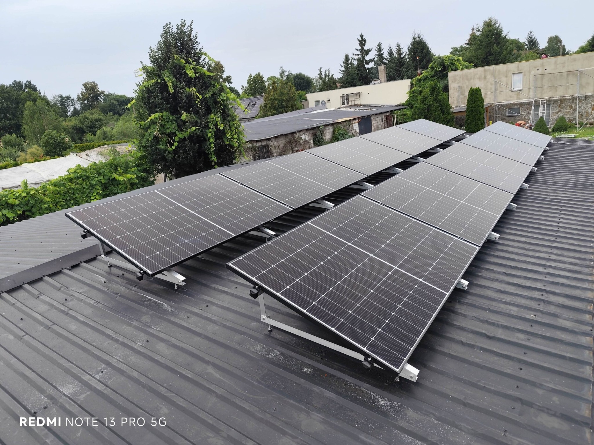 Rząd paneli fotowoltaicznych na ciemnym, metalowym dachu budynku. Widok z góry, w tle zieleń drzew i zabudowania. Montaż na srebrnych wspornikach.