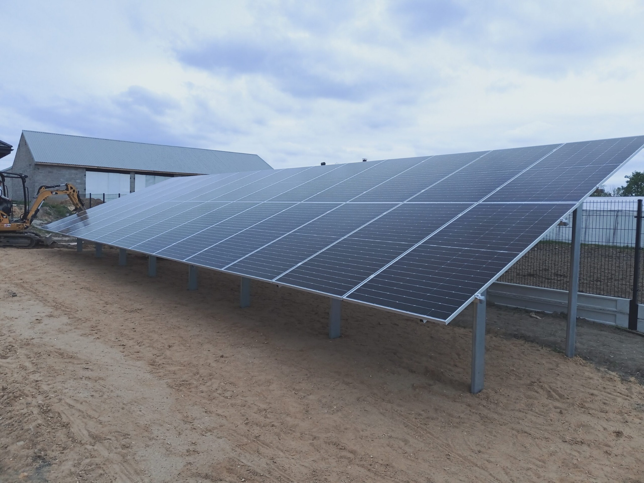Instalacja fotowoltaiczna na gruncie, panele zamontowane na metalowych słupkach, w tle budynek gospodarczy i ogrodzenie. Widok z dołu, pochmurne niebo.