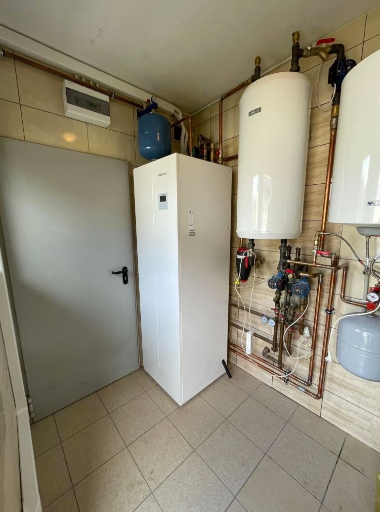 Biała pompa ciepła marki Kaisai w pomieszczeniu z beżowymi płytkami, obok szare drzwi i zbiorniki wodne z miedzianymi rurami. Widok z niskiego kąta.
