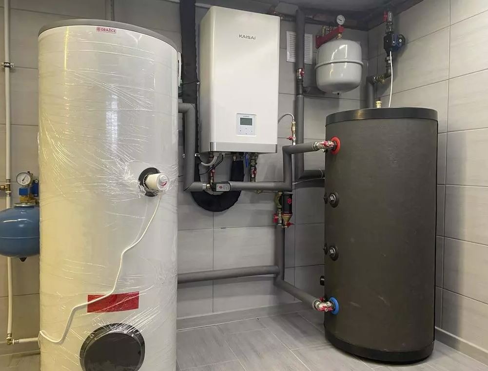Kotłownia z dwoma zbiornikami, białym kotłem Kaisai i szarymi rurami, całość w pomieszczeniu wyłożonym jasnymi płytkami. Jeden zbiornik owinięty folią.