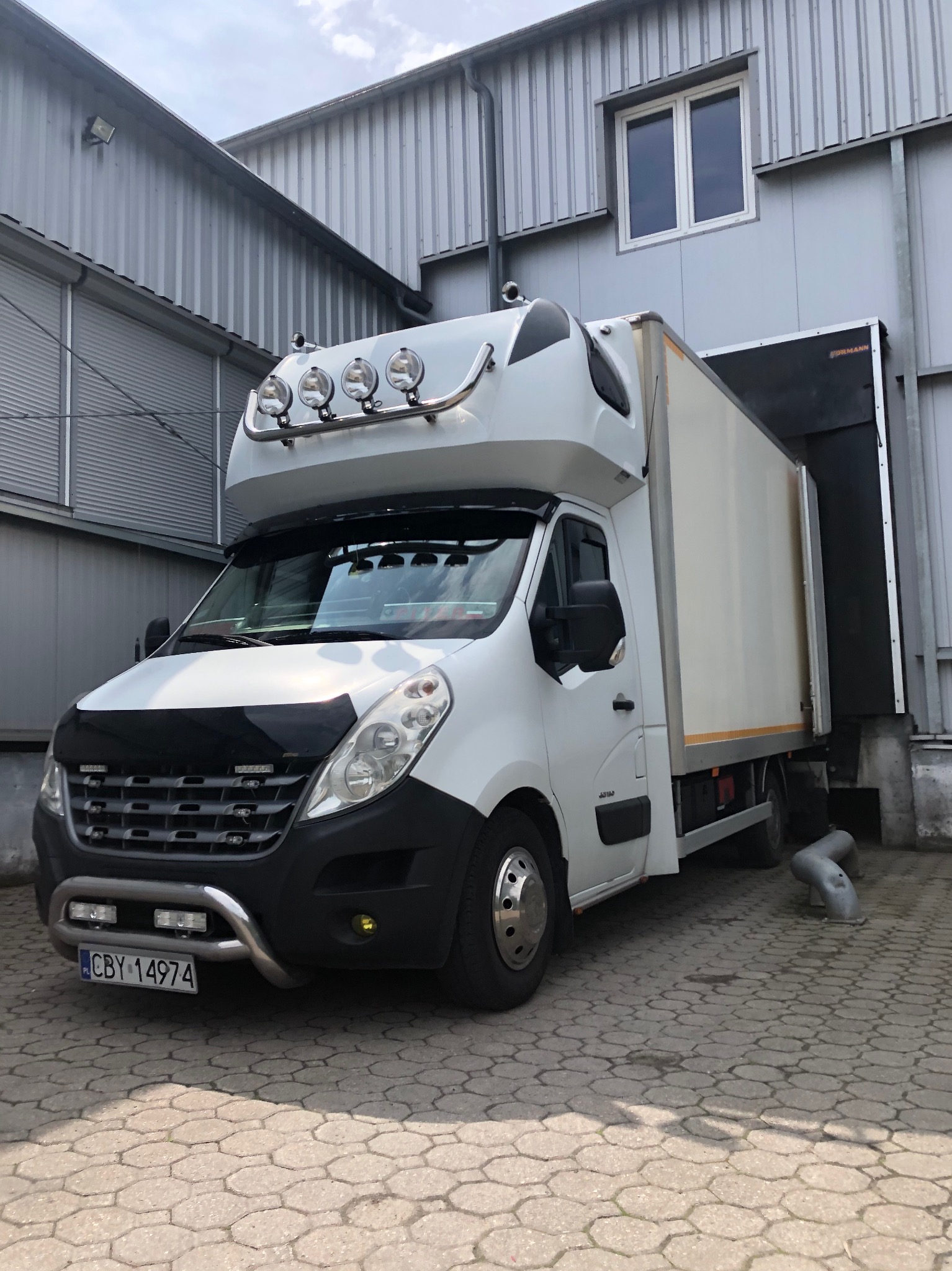 Biały bus dostawczy z dodatkowym oświetleniem na dachu, zaparkowany przy rampie załadunkowej z szarej blachy falistej.