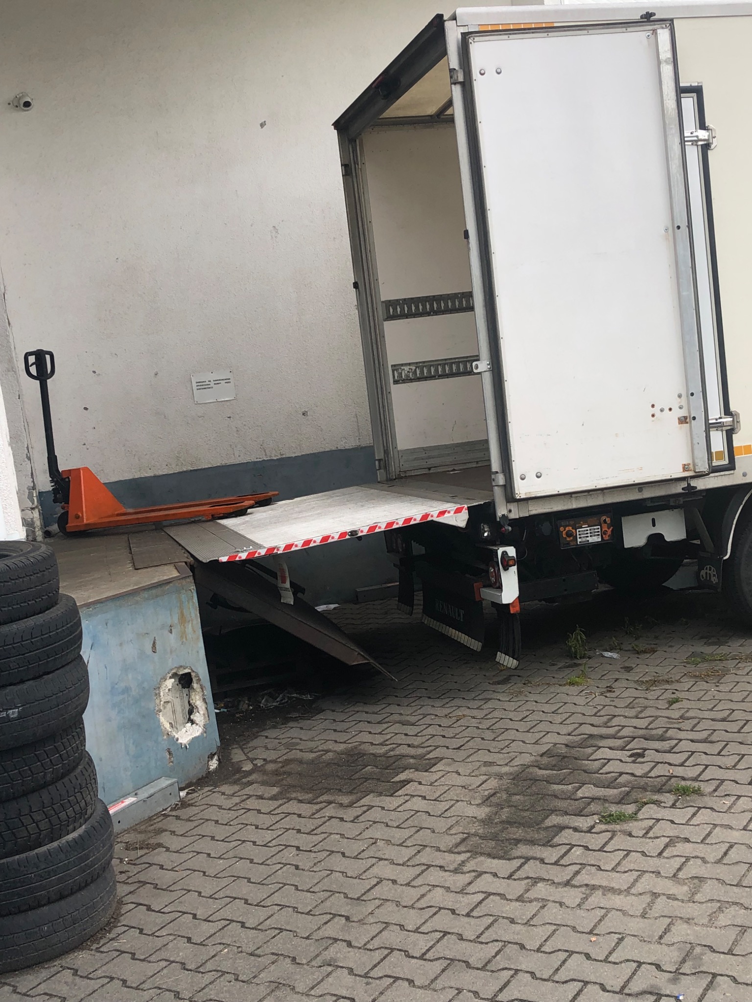 Otwarty biały furgon dostawczy z rampą załadunkową, obok stos opon i wózka paletowego.