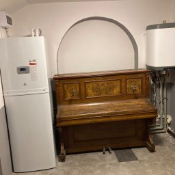 Niedźwiedź-Sanit Sp.J. - Biała pompa ciepła zamontowana w piwnicy obok starego pianina, widoczny również bojler elektryczny i instalacja grzewcza na ścianie.