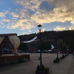 Niedźwiedź-Sanit Sp.J. - Widok na plac miejski z oświetleniem ulicznym w stylu retro, w tle zalesione wzgórze z częściowo ośnieżonym stokiem narciarskim i skocznią narciarską, pochmurne niebo o zachodzie słońca.