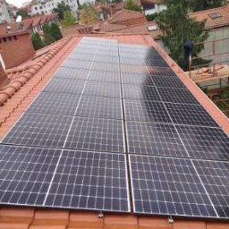 Niedźwiedź-Sanit Sp.J. - Panele słoneczne zamontowane na dachu z czerwonej dachówki, widoczne elementy mocujące i fragment sylwetki osoby pracującej przy instalacji, w tle zabudowa miejska.