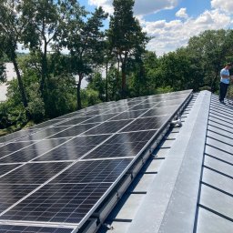 Niedźwiedź-Sanit Sp.J. - Instalacja paneli fotowoltaicznych na metalowym dachu z widokiem na jezioro i las, pracownik w niebieskiej koszulce sprawdza coś na telefonie.