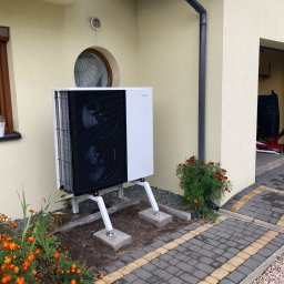 Niedźwiedź-Sanit Sp.J. - Biała pompa ciepła zamontowana na zewnątrz budynku z beżową elewacją, widoczne kwiaty i kostka brukowa.