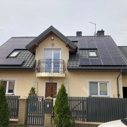 Niedźwiedź-Sanit Sp.J. - Dom jednorodzinny z zamontowanymi panelami fotowoltaicznymi na dachu pokrytym dachówką, widoczny balkon z ozdobną balustradą i adres 'ul. Debowa 12' na ogrodzeniu.