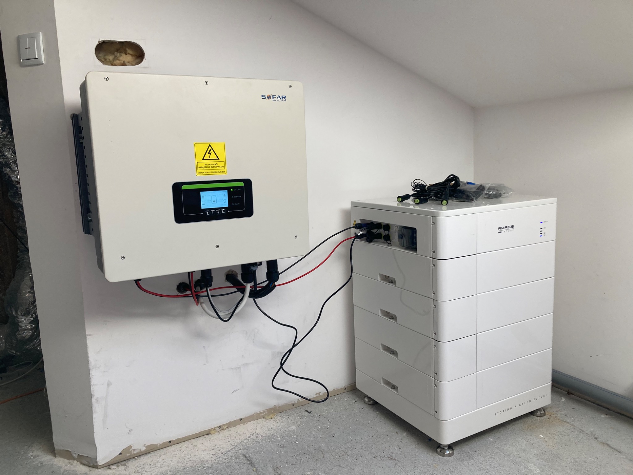Biały inwerter SOFAR przymocowany do ściany, połączony przewodami z modułową baterią magazynującą energię AMASS, widoczne niedoskonałości wykończenia ściany.