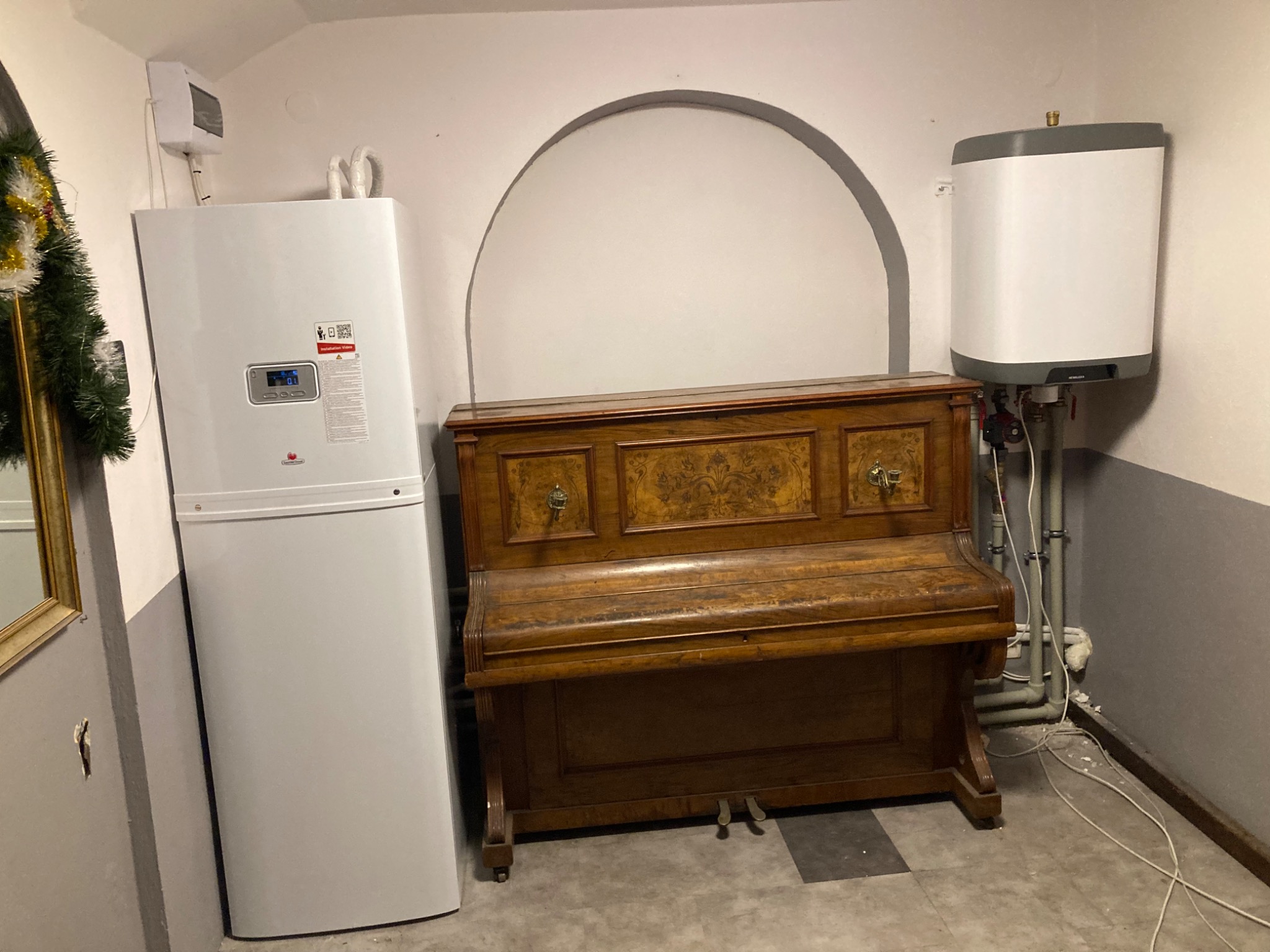 Biała pompa ciepła zamontowana w piwnicy obok starego pianina, widoczny również bojler elektryczny i instalacja grzewcza na ścianie.