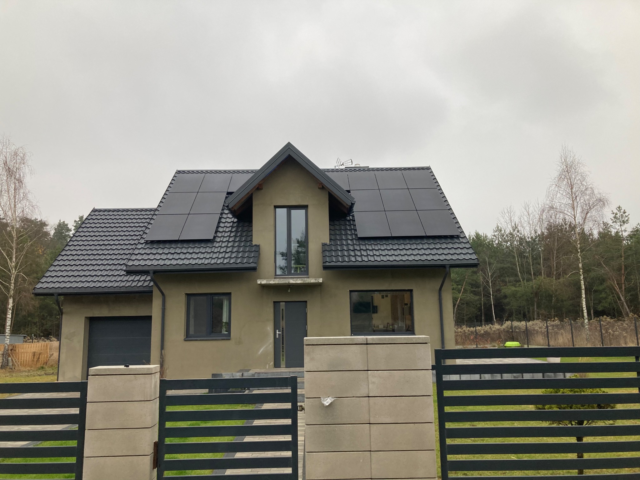 Dom jednorodzinny z szarym dachem i panelami fotowoltaicznymi na dachu, widok zza nowoczesnego, metalowego ogrodzenia w pochmurny dzień.