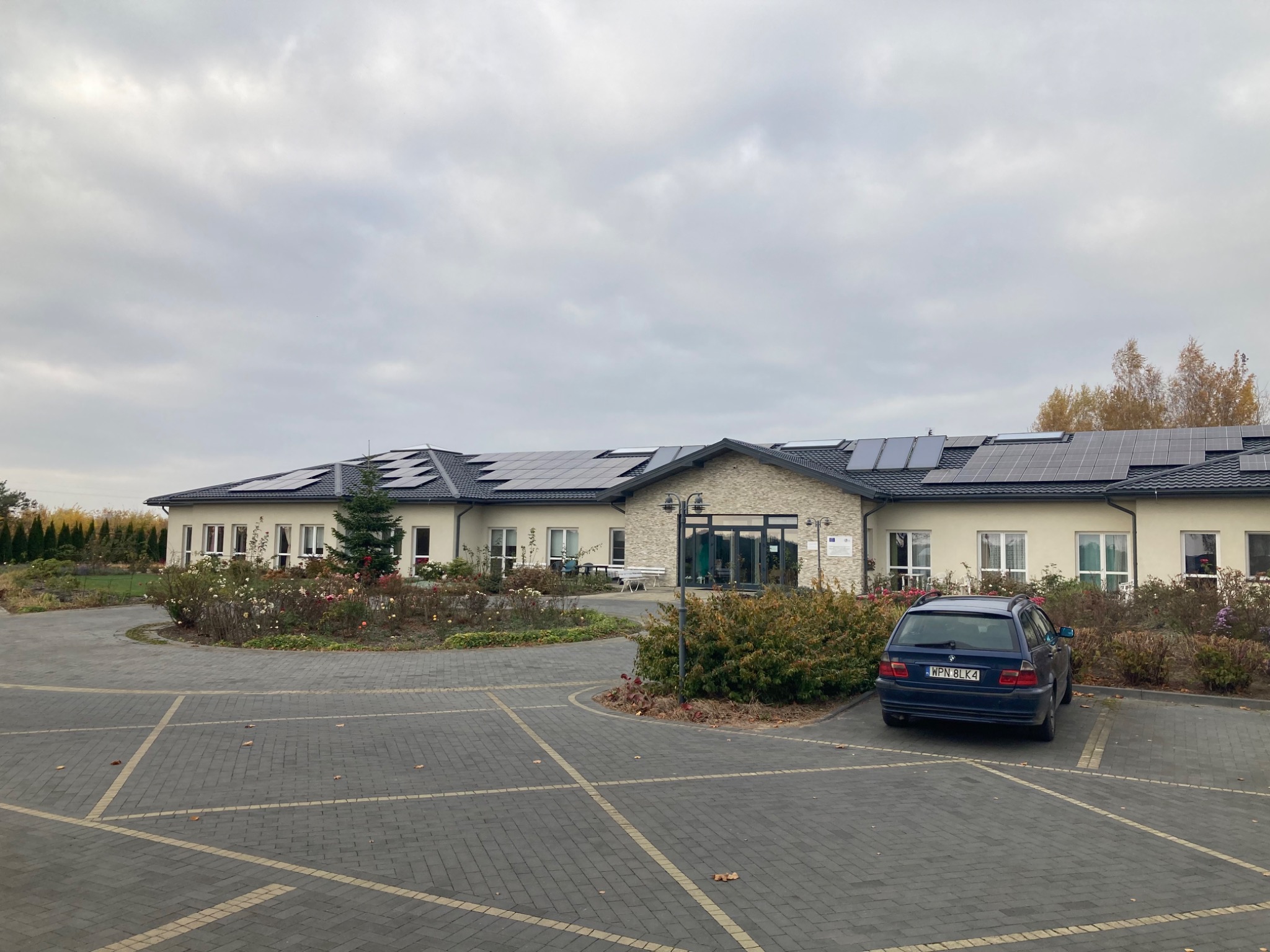 Budynek z panelami fotowoltaicznymi na dachu, widoczny z parkingu w Nowym Dworze Mazowieckim, z zaparkowanym samochodem osobowym z przodu. Pochmurne niebo w tle.