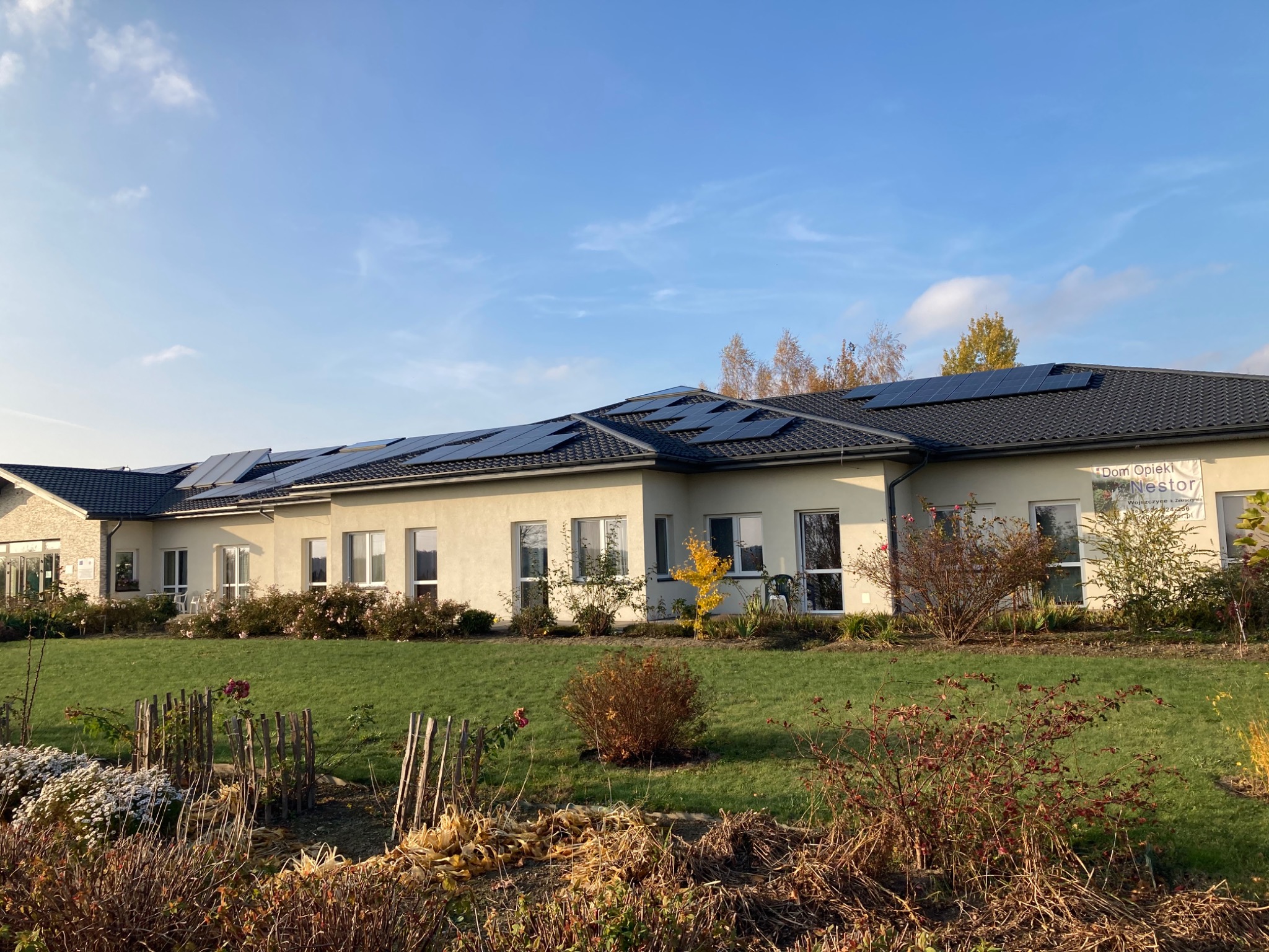 Budynek Domu Opieki Nestor z panelami fotowoltaicznymi na dachu, otoczony trawnikiem i krzewami w jesiennej scenerii.
