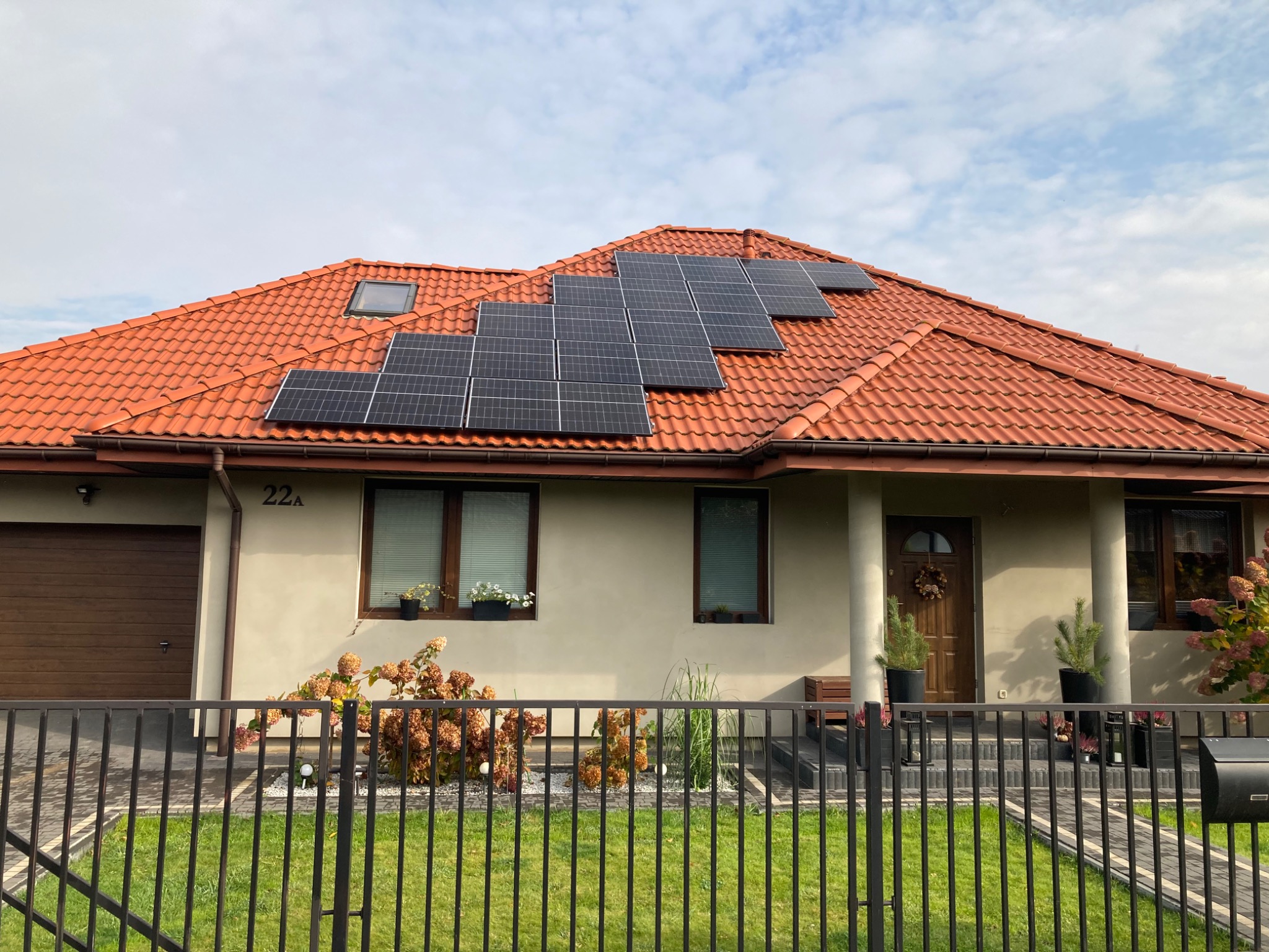 Czarny panel fotowoltaiczny na pomarańczowym dachu domu jednorodzinnego z numerem 22A, widziany zza czarnego ogrodzenia.