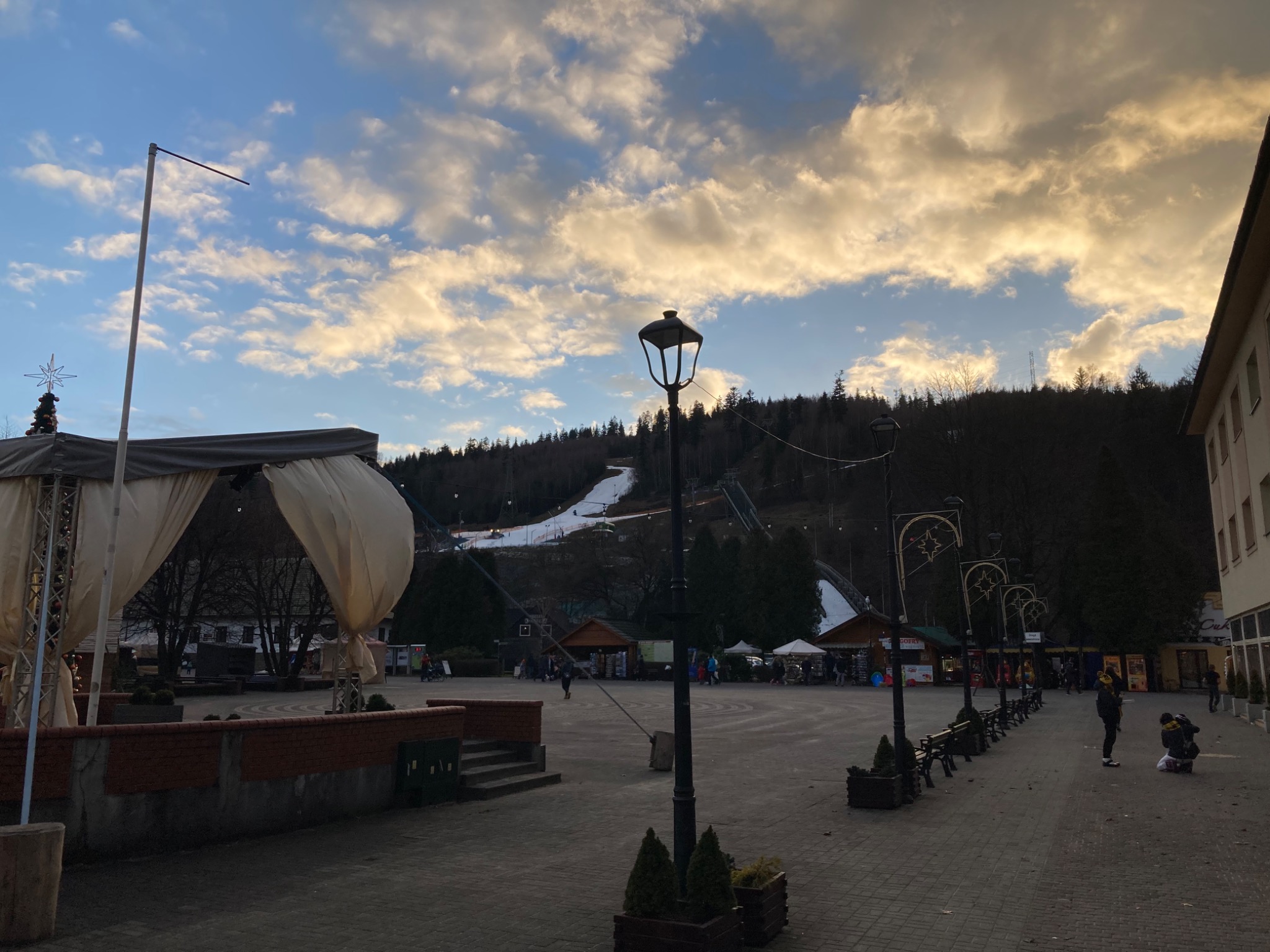 Widok na plac miejski z oświetleniem ulicznym w stylu retro, w tle zalesione wzgórze z częściowo ośnieżonym stokiem narciarskim i skocznią narciarską, pochmurne niebo o zachodzie słońca.