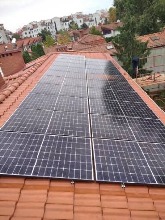 Panele słoneczne zamontowane na dachu z czerwonej dachówki, widoczne elementy mocujące i fragment sylwetki osoby pracującej przy instalacji, w tle zabudowa miejska.