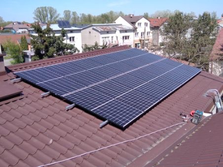 Panele fotowoltaiczne zamontowane na brązowym dachu domu jednorodzinnego, widoczne narzędzia i drabina, w tle zabudowa miejska i błękitne niebo.