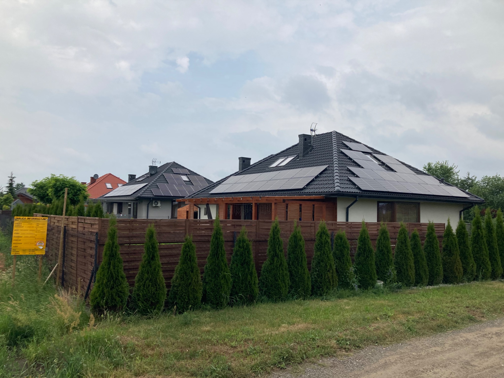 Dwa domy jednorodzinne z panelami słonecznymi na dachu, oddzielone drewnianym ogrodzeniem z tujami, tablica informacyjna po lewej stronie.