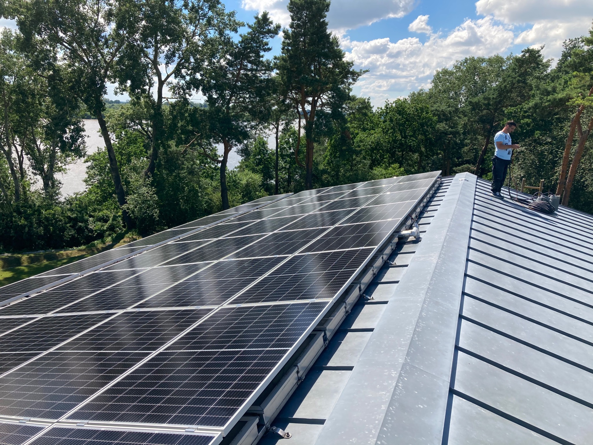Instalacja paneli fotowoltaicznych na metalowym dachu z widokiem na jezioro i las, pracownik w niebieskiej koszulce sprawdza coś na telefonie.