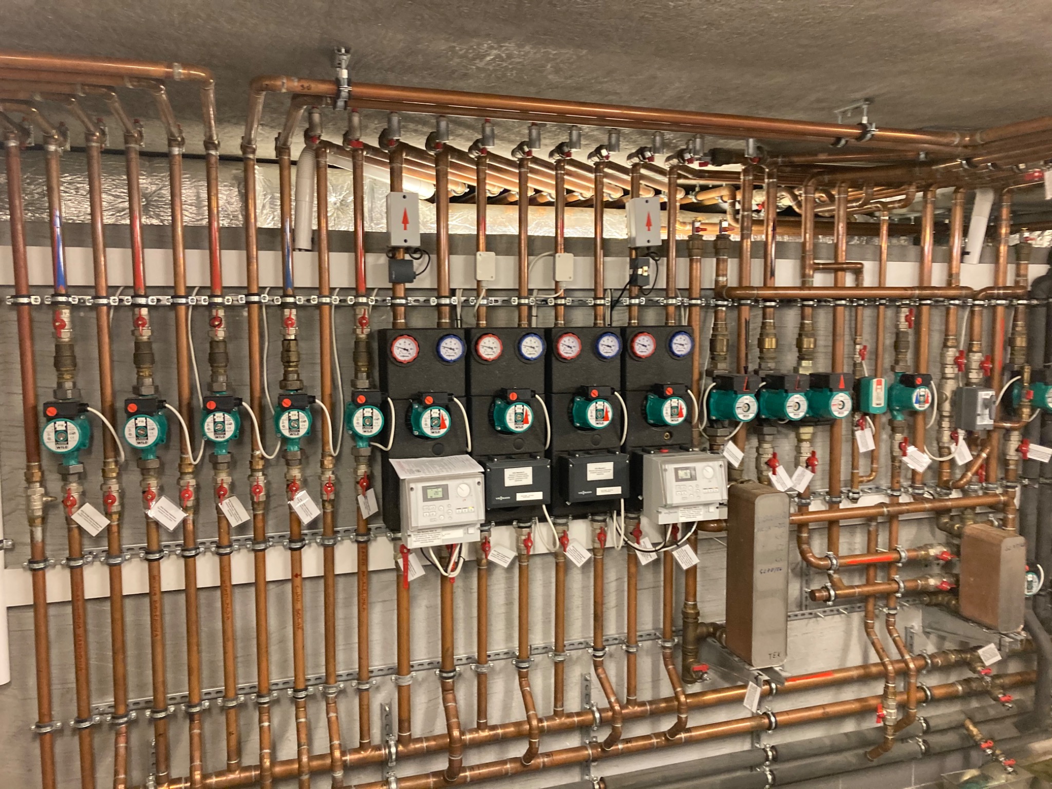 Złożona instalacja z miedzianych rur, zaworów, pomp obiegowych WILO, manometrów i sterowników zamontowana na ścianie w kotłowni.