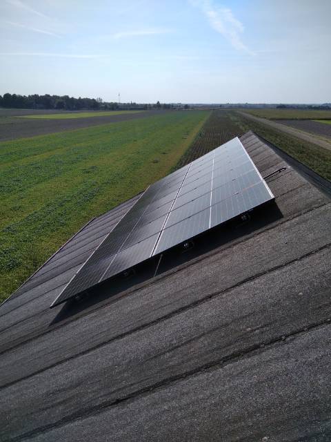 Panel fotowoltaiczny zamontowany na dachu z papy, widok z góry, w tle pola uprawne i błękitne niebo.