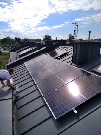 Montaż paneli słonecznych na dachu z blachy, widoczne odbicie słońca na panelach, instalator w trakcie pracy.