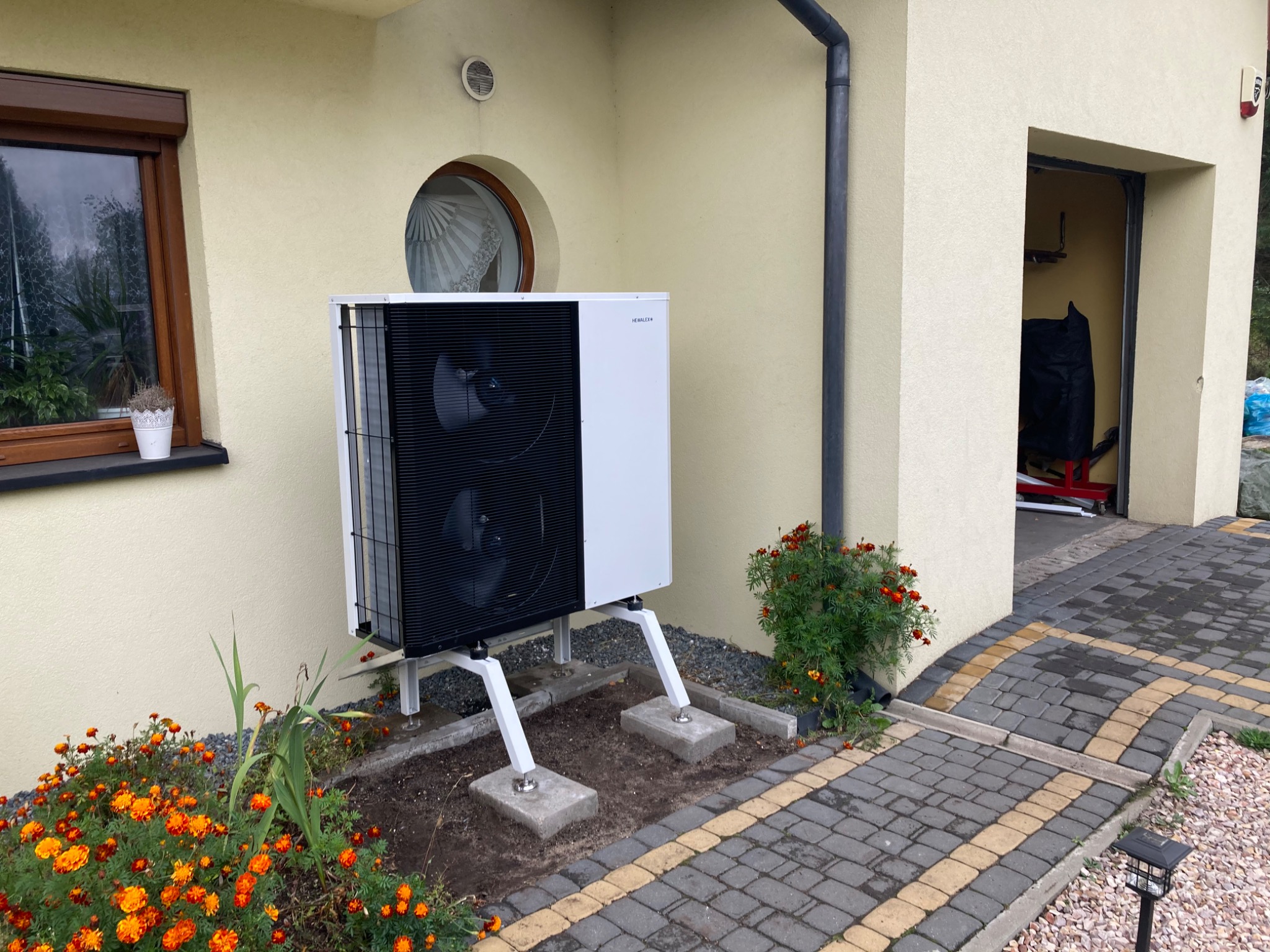 Biała pompa ciepła zamontowana na zewnątrz budynku z beżową elewacją, widoczne kwiaty i kostka brukowa.