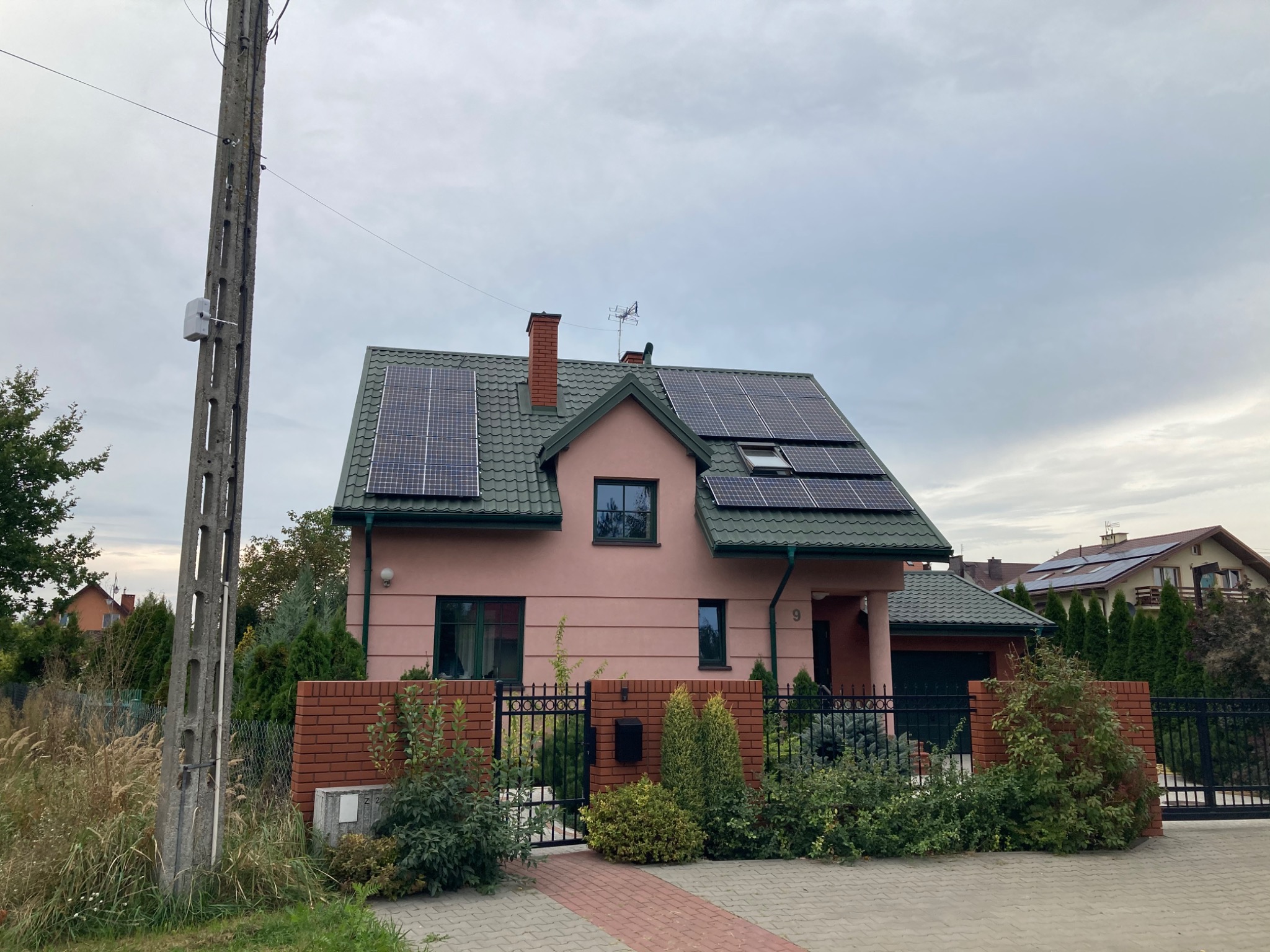 Różowy dom jednorodzinny z zielonym dachem i panelami słonecznymi, widok z ulicy w pochmurny dzień.