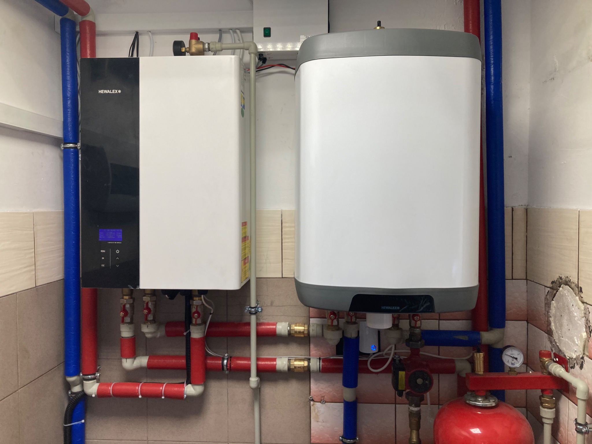 Zestaw urządzeń grzewczych z widocznymi rurami w kolorach niebieskim i czerwonym, zamontowany na ścianie wyłożonej płytkami. Widoczny manometr i czerwony zbiornik wyrównawczy.