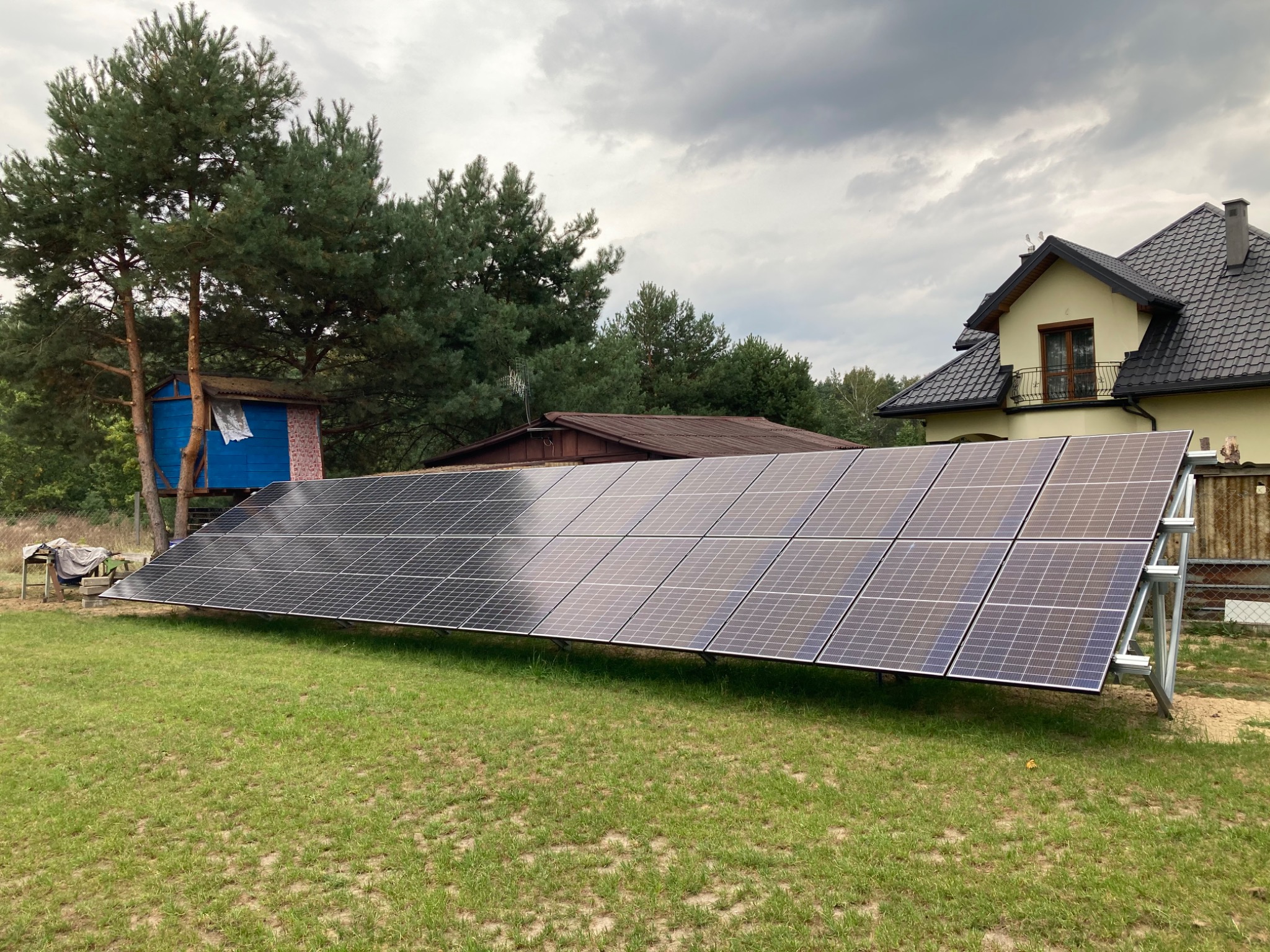 Rząd paneli fotowoltaicznych na metalowej konstrukcji, trawnik, w tle dom z ciemnym dachem i budka na drzewie, pochmurne niebo.