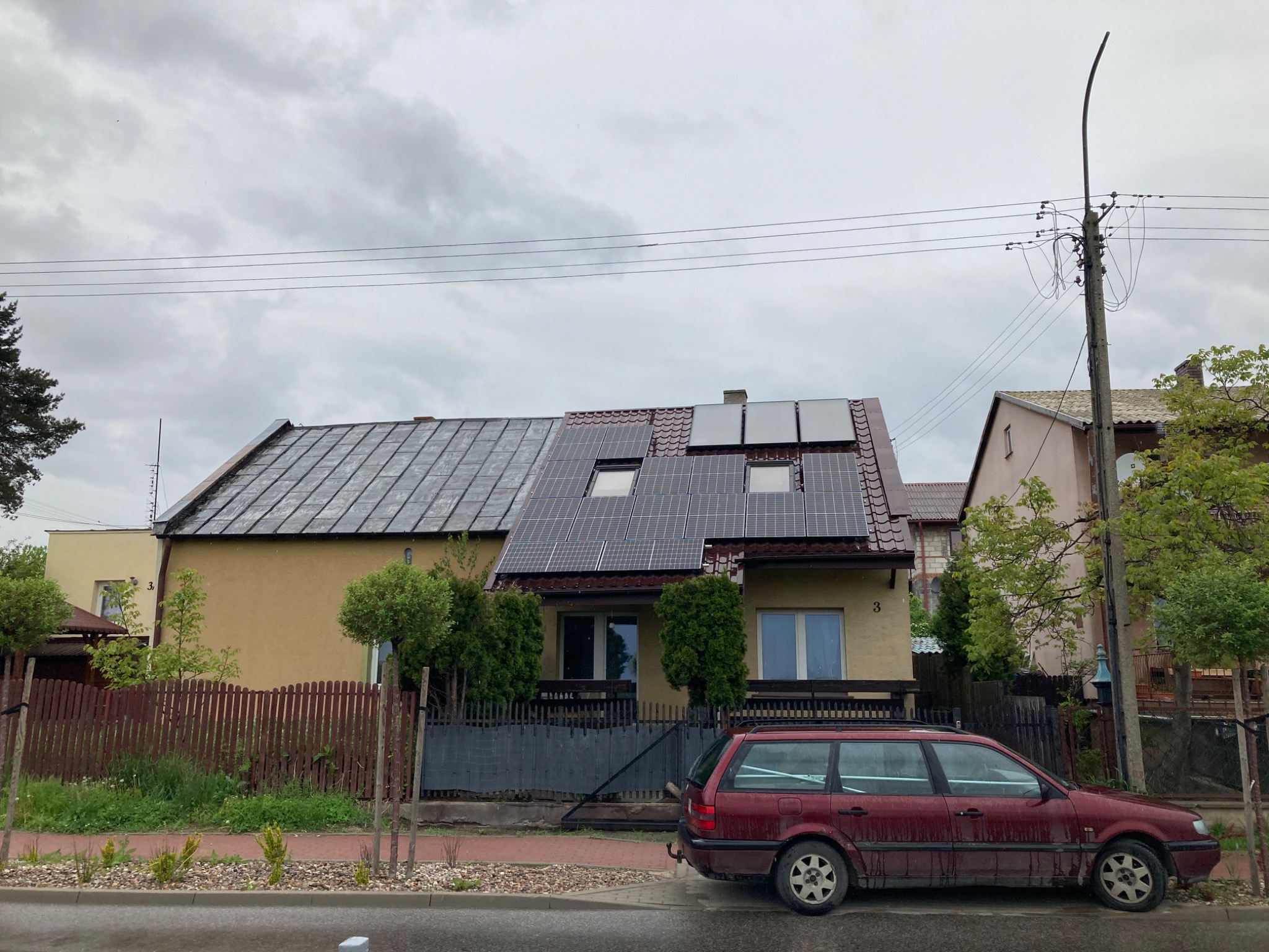 Dom jednorodzinny z panelami słonecznymi na dachu, widok z ulicy w pochmurny dzień, z zaparkowanym bordowym kombi