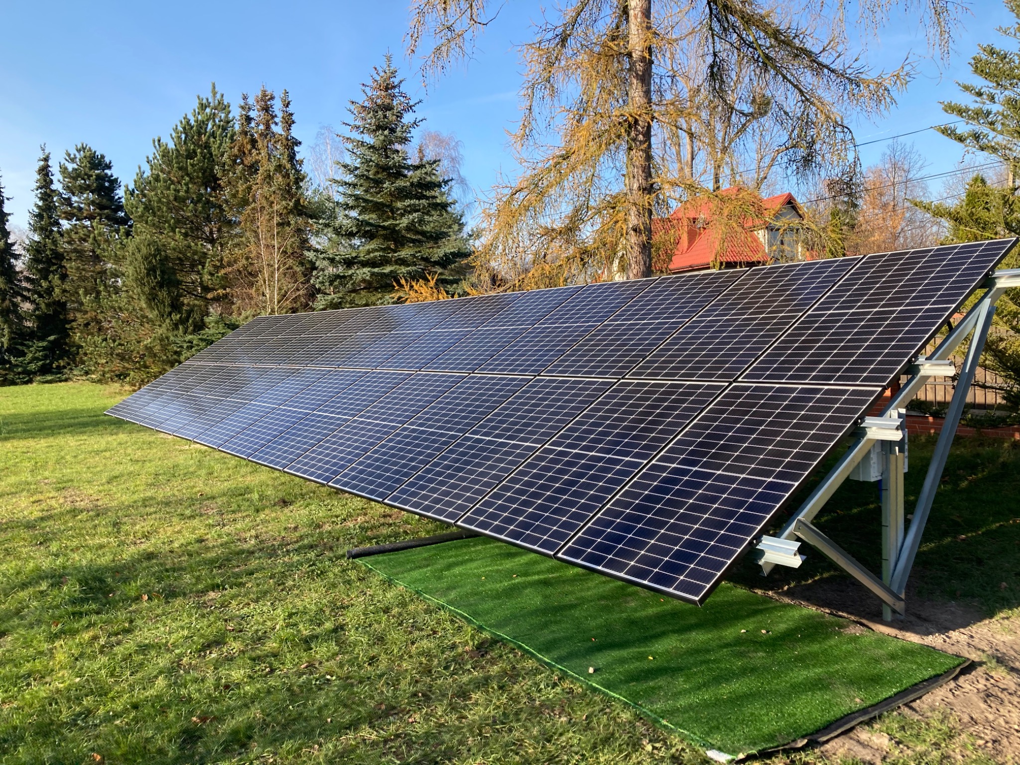 Rząd paneli fotowoltaicznych na metalowej konstrukcji, zamontowany na trawniku z fragmentem sztucznej trawy, w tle drzewa i budynek z czerwonym dachem.