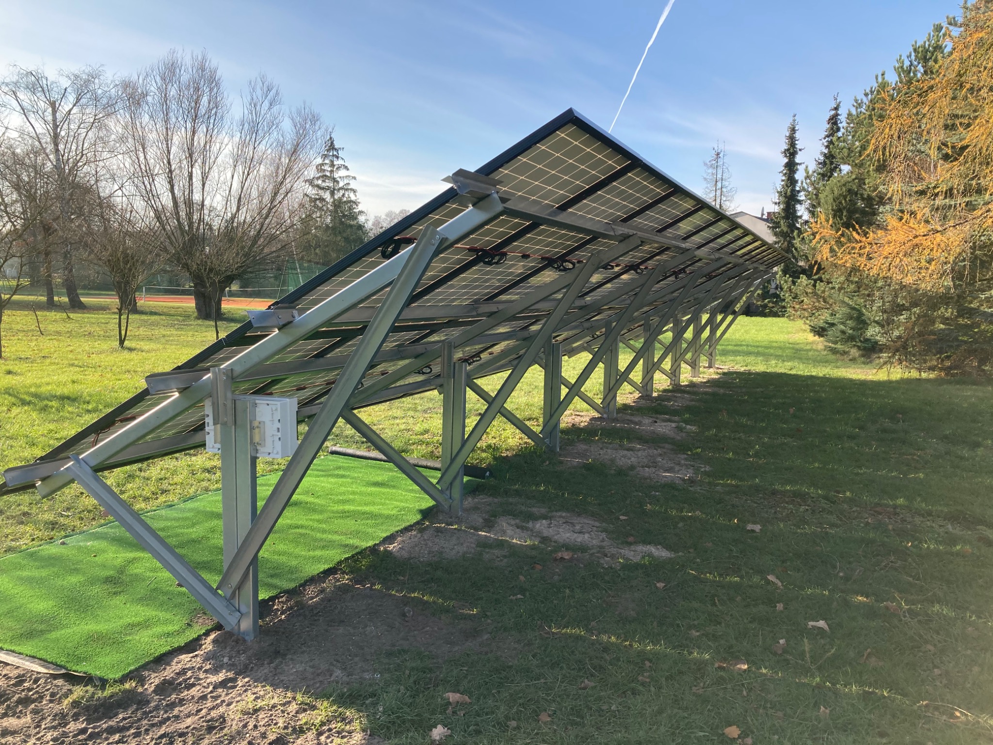 Rząd paneli fotowoltaicznych na metalowej konstrukcji ustawiony na trawie, widok od spodu z widocznymi mocowaniami i elementami instalacji elektrycznej, tło stanowią drzewa i błękitne niebo.