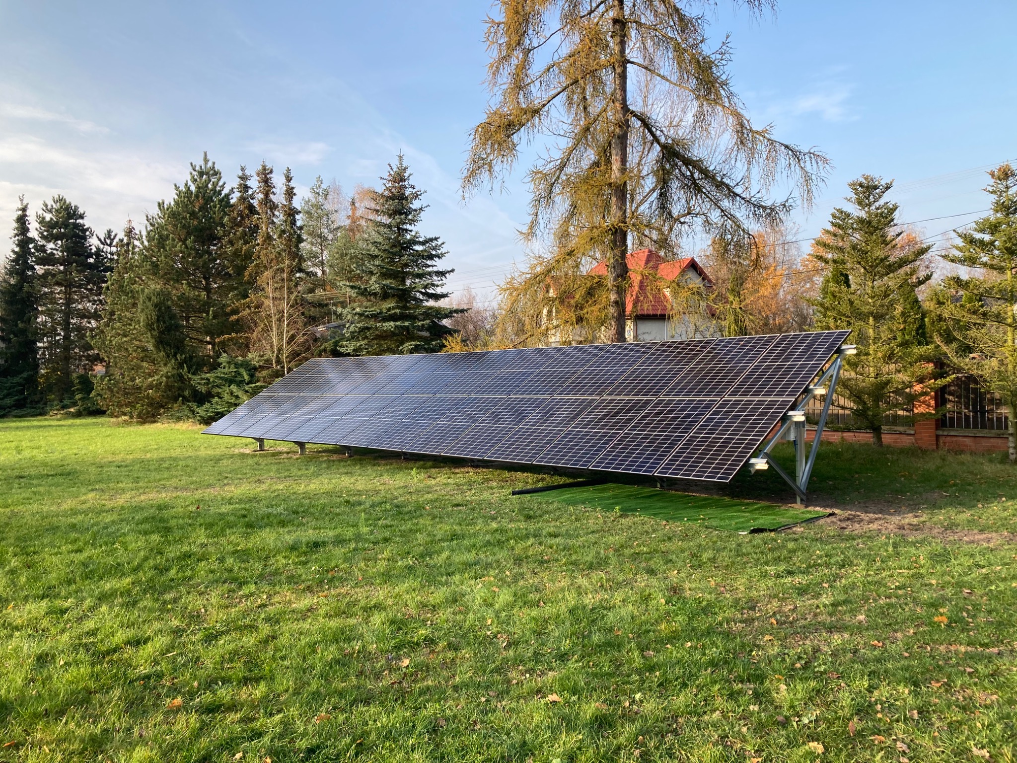 Rząd paneli słonecznych na metalowej konstrukcji, umieszczony na trawniku przed domem z czerwonym dachem, otoczony drzewami w jesiennej scenerii.