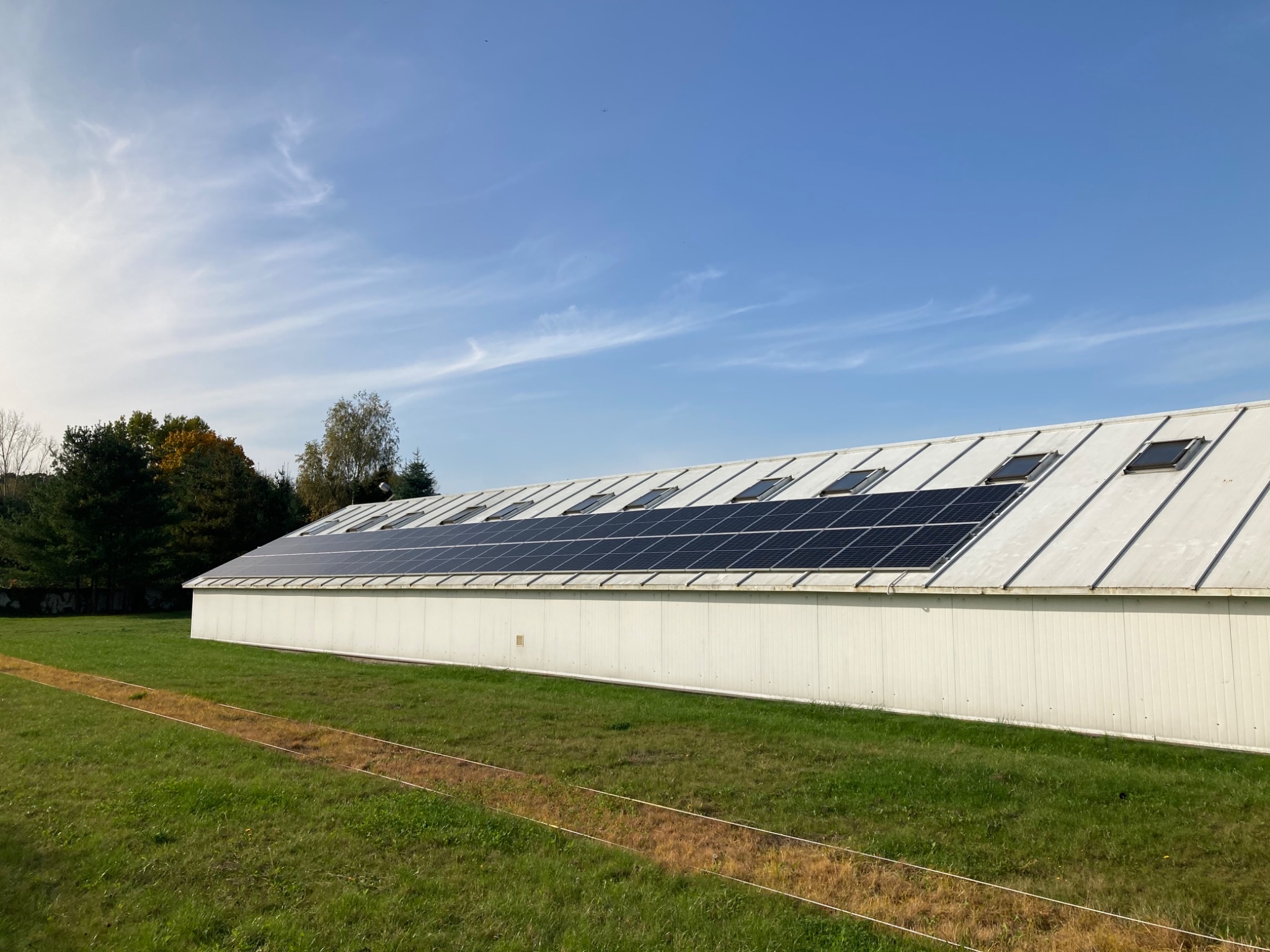 Duży budynek gospodarczy z białymi ścianami i panelami słonecznymi na dachu, widoczne okna dachowe, trawnik z brązowymi śladami na pierwszym planie, błękitne niebo z delikatnymi chmurami w tle.