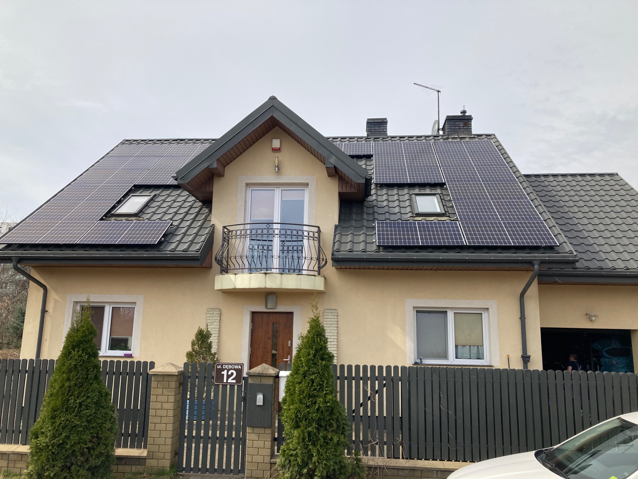 Dom jednorodzinny z zamontowanymi panelami fotowoltaicznymi na dachu pokrytym dachówką, widoczny balkon z ozdobną balustradą i adres 'ul. Debowa 12' na ogrodzeniu.