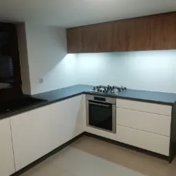 Nowoczesna kuchnia w kształcie litery L z białymi frontami szafek i blatem w kolorze grafitowym, oświetlona listwą LED pod górnymi szafkami z drewnopodobnym wykończeniem; widoczny zlew i płyta gazowa.